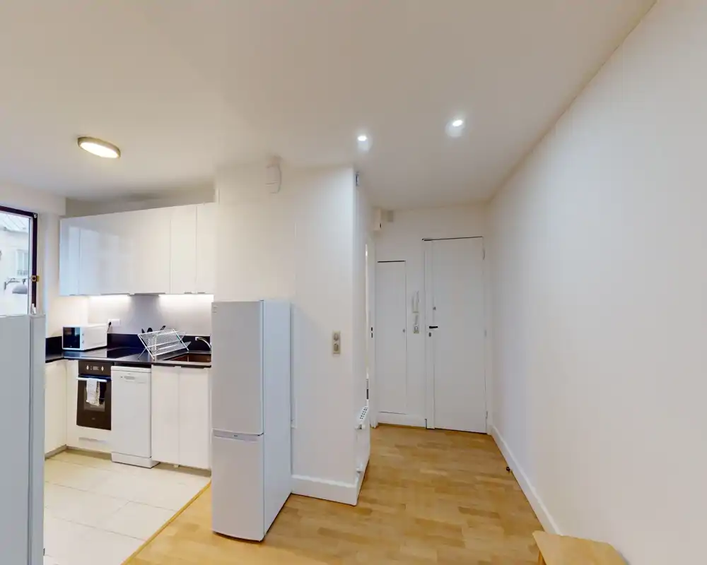 Compact open-plan kitchenette with modern white cabinets, integrated oven and dishwasher, and a freestanding fridge. Bright, clean and practical for a small apartment.