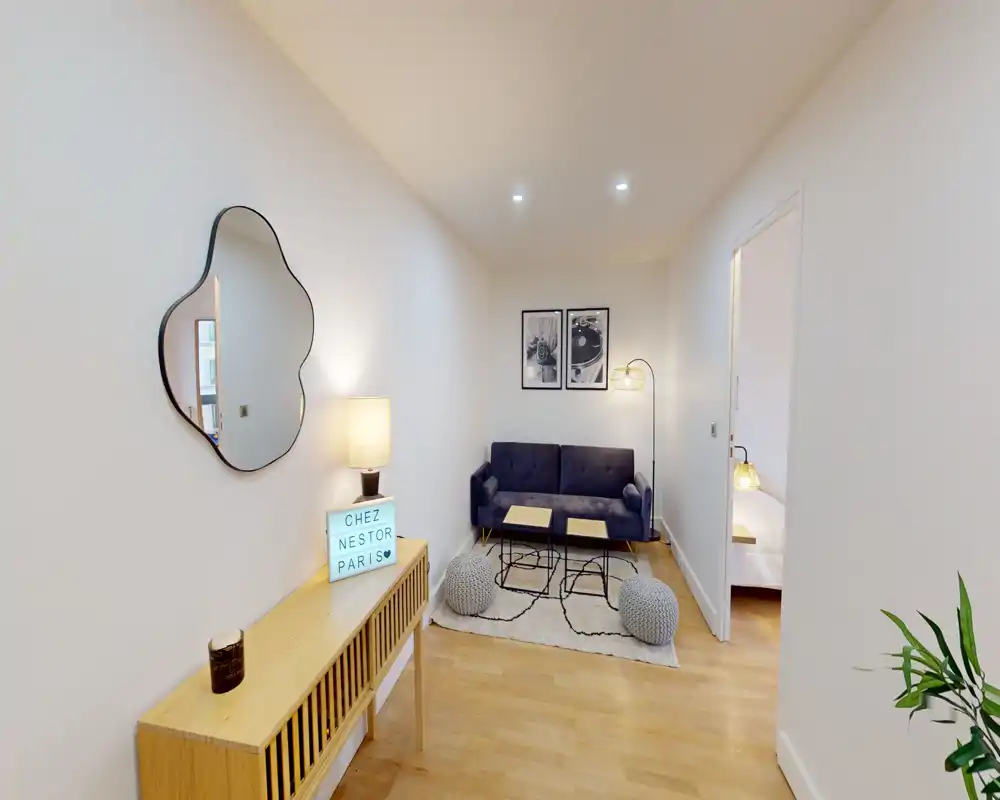 Small stylish living area with a compact sofa, coffee tables, poufs and warm lighting — bright white walls and wooden floor create a welcoming atmosphere.