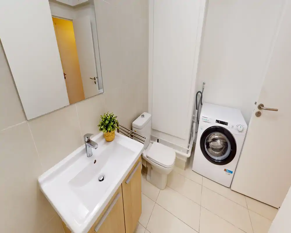 Compact, modern bathroom with a sink and mirror, toilet and a front-loading washing machine; light tiles and wooden cabinet give a clean, functional look.