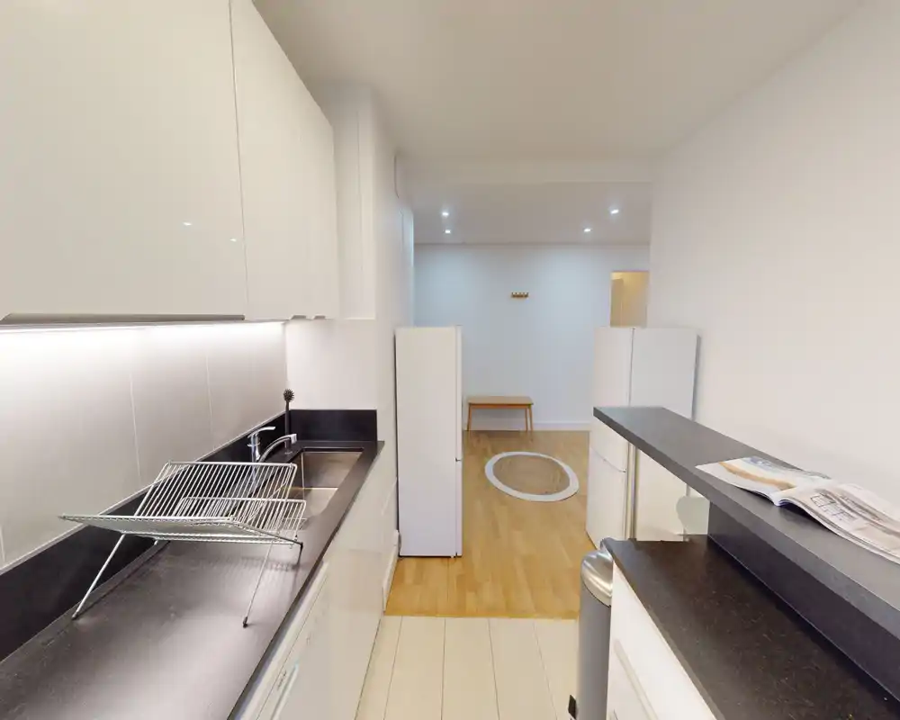 Modern compact kitchen with black countertops, white cabinetry and integrated sink; opens onto a small dining or living area with wooden floors.