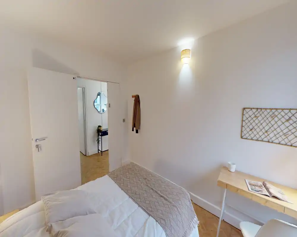 Bright, simply styled bedroom with a neatly made bed, a small desk and soft wall lighting—ideal for a cozy single or couple.