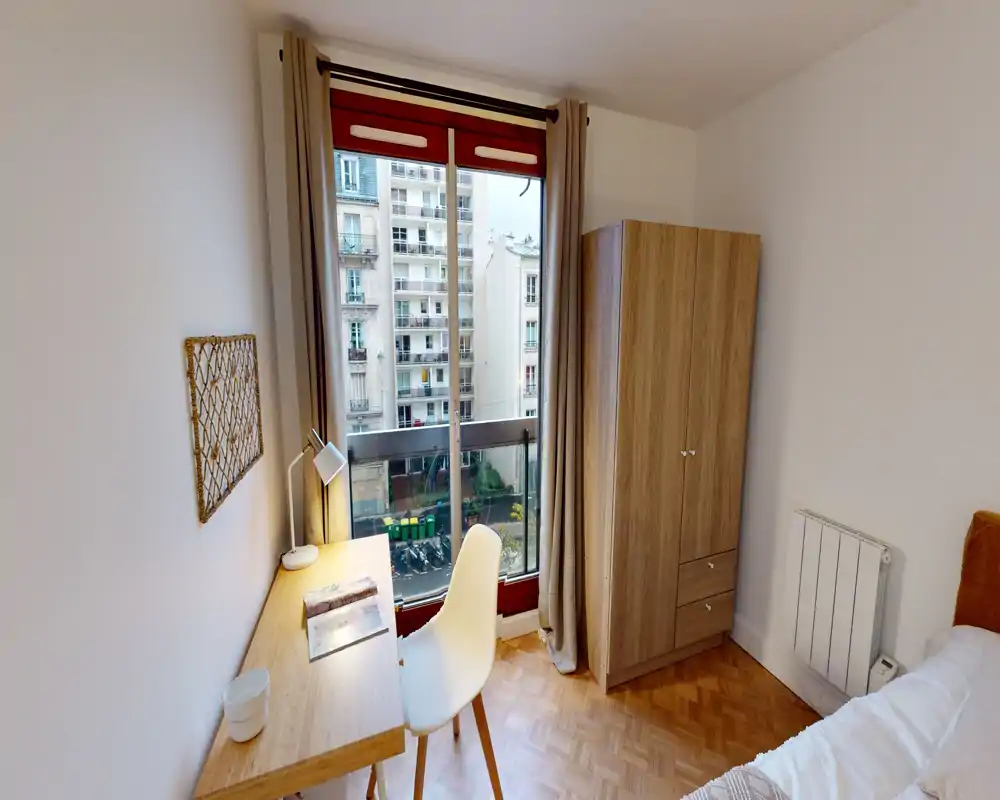 Bright, tidy bedroom corner with a study desk by a large window, a wardrobe and a glimpse of the bed — ideal for a single occupant or student.