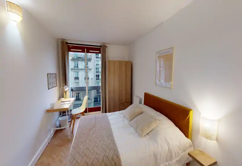 Bright, neatly styled bedroom with a double bed, cozy textiles and a small desk by the window. Natural light from the large window and warm bedside lamps create a welcoming atmosphere.