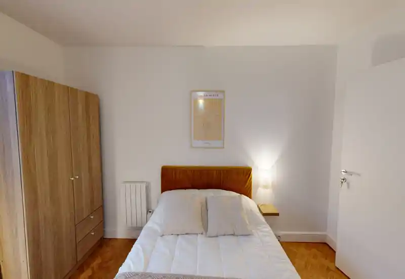 Neat, well-lit bedroom with a comfortable double bed, warm headboard and a wooden wardrobe. Soft bedding and a bedside lamp create a cozy atmosphere ideal for rental photos.