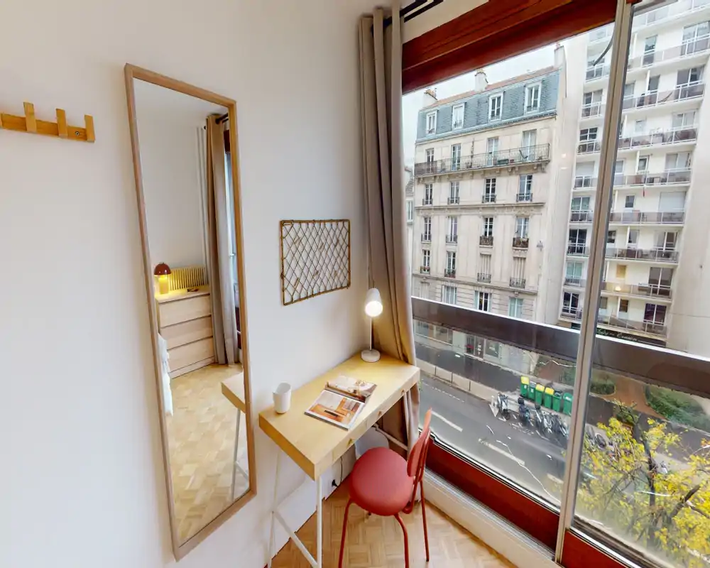 Coin de chambre compact avec un petit bureau près d'une grande fenêtre donnant sur la rue ; un miroir pleine longueur reflète une autre partie de la zone nuit. Mobilier lumineux et minimaliste de style scandinave, parfait pour un voyageur seul.