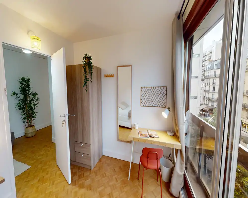 Espace chambre lumineux et chaleureux avec parquet, armoire et petit bureau près d'une grande fenêtre donnant sur la ville ; une partie du lit est visible par la porte ouverte.