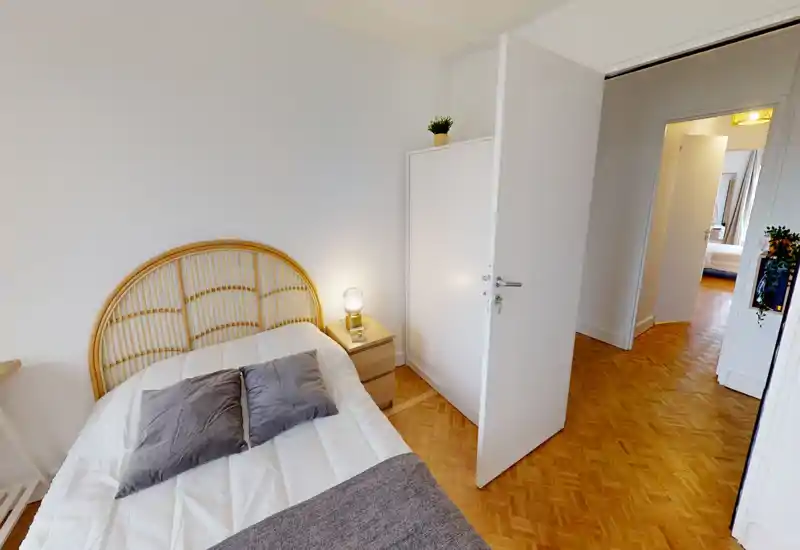 Bright, cozy single bedroom with a rattan headboard, bedside table and lamp; parquet flooring and clean white walls create a calm, welcoming space.