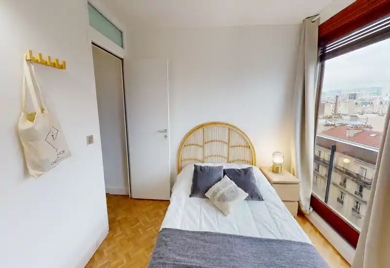 Bright, well-styled bedroom with a rattan headboard, decorative cushions and a large window offering city views.