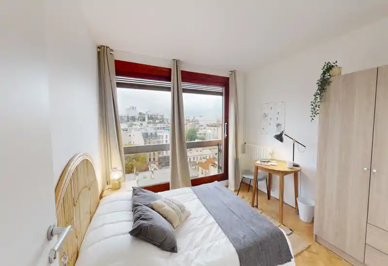 Bright, cozy bedroom with a large window offering a city view, a neatly made double bed with decorative pillows and a small desk by the wardrobe — ideal for single or couple stays.