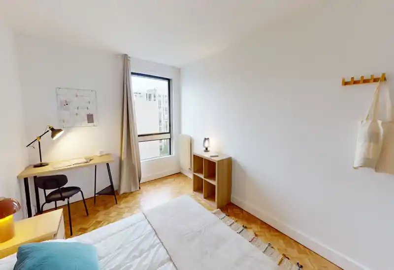 Camera da letto luminosa e minimal con letto ben visibile, comodino e una scrivania vicino alla finestra. La luce naturale e il pavimento in legno creano un’atmosfera accogliente perfetta per annunci di affitto.