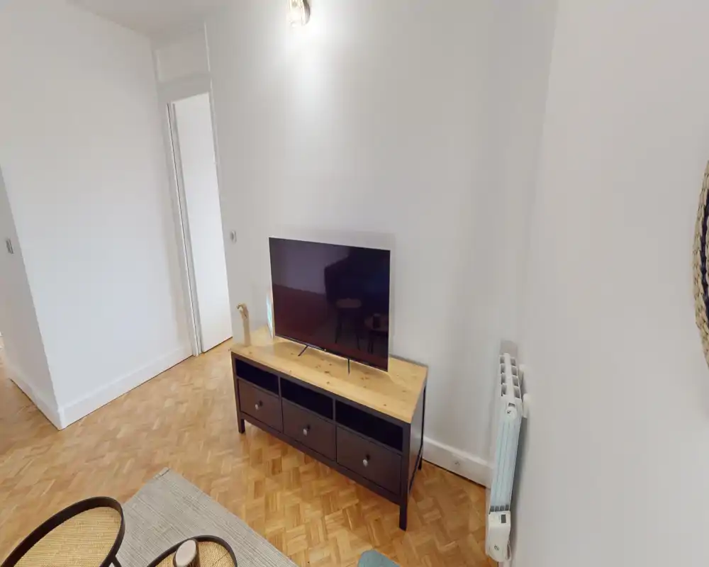 Simple, well-lit living area with a TV on a console, parquet flooring and a neutral, minimal decor—suitable for a compact lounge or studio setup.