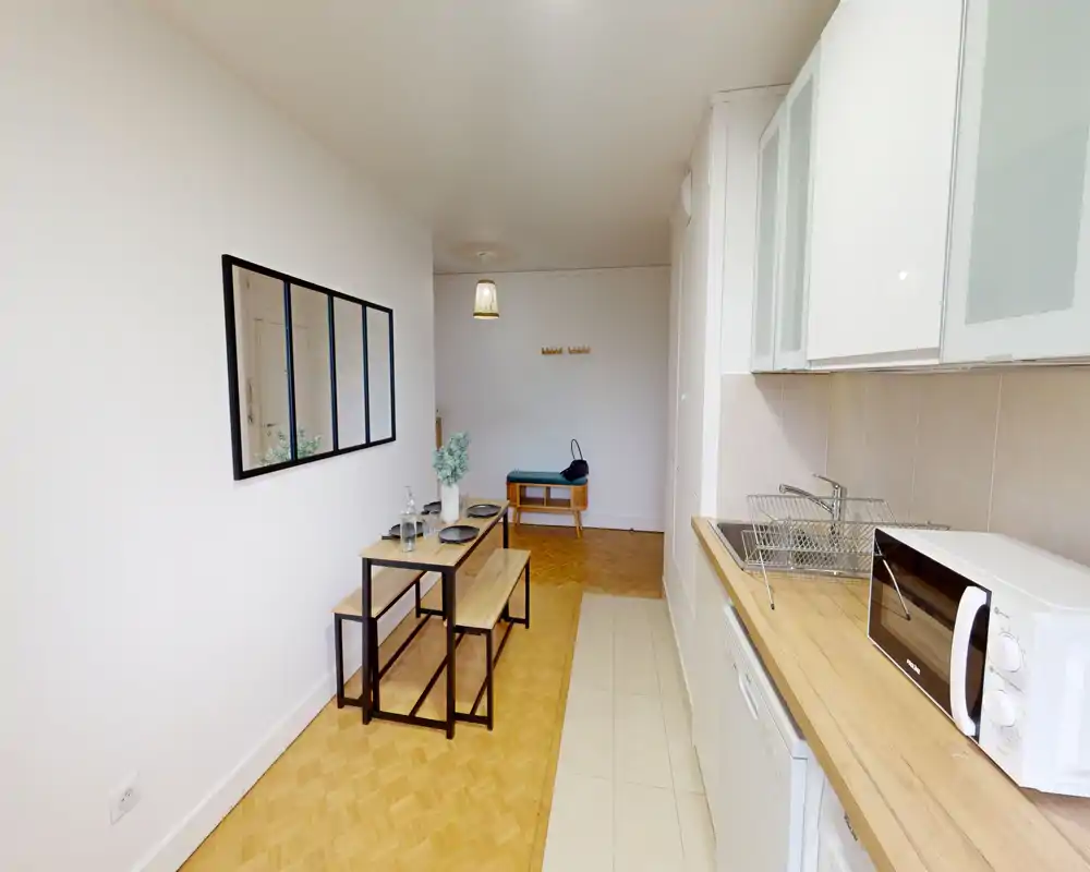 Well-lit compact kitchen with a wooden countertop, microwave and sink; a small dining bench setup and decorative plant create a functional, cozy space ideal for a studio apartment.