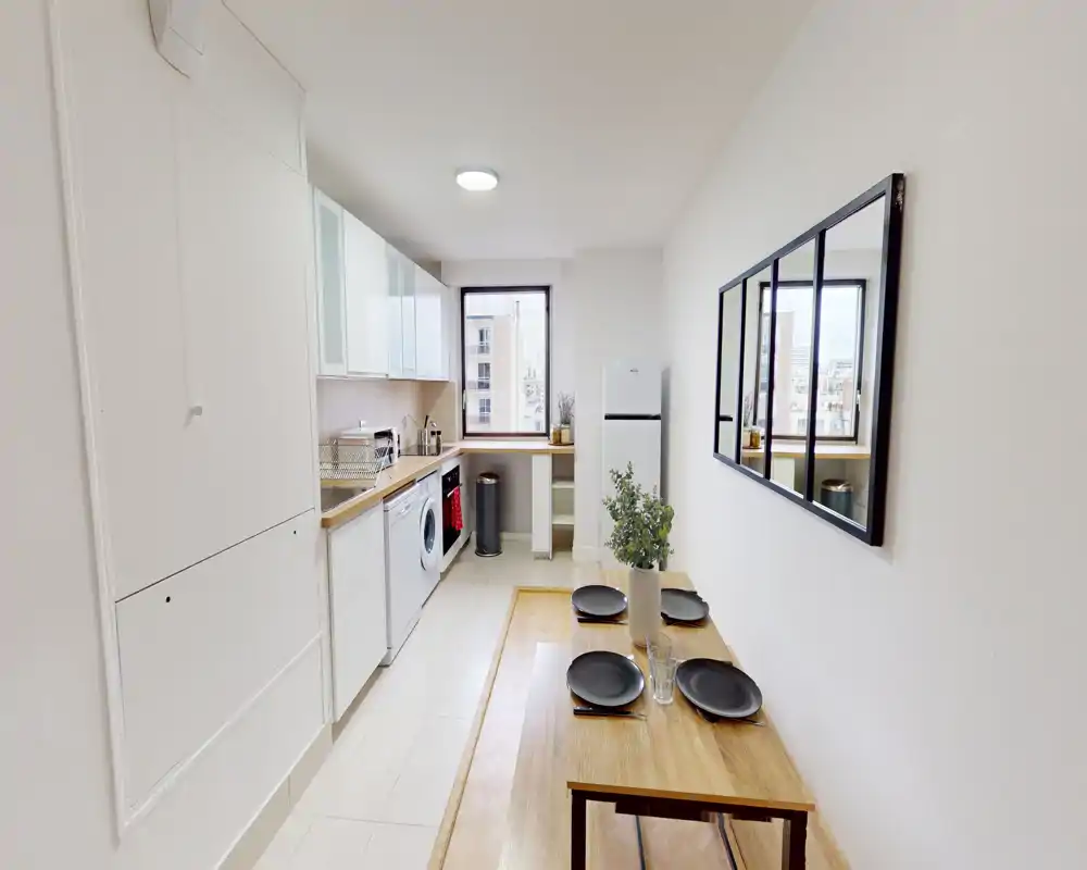 Bright, modern galley kitchen with white cabinets, wooden countertops and a small dining table set for four; appliances include a washing machine and fridge, with a window providing natural light.