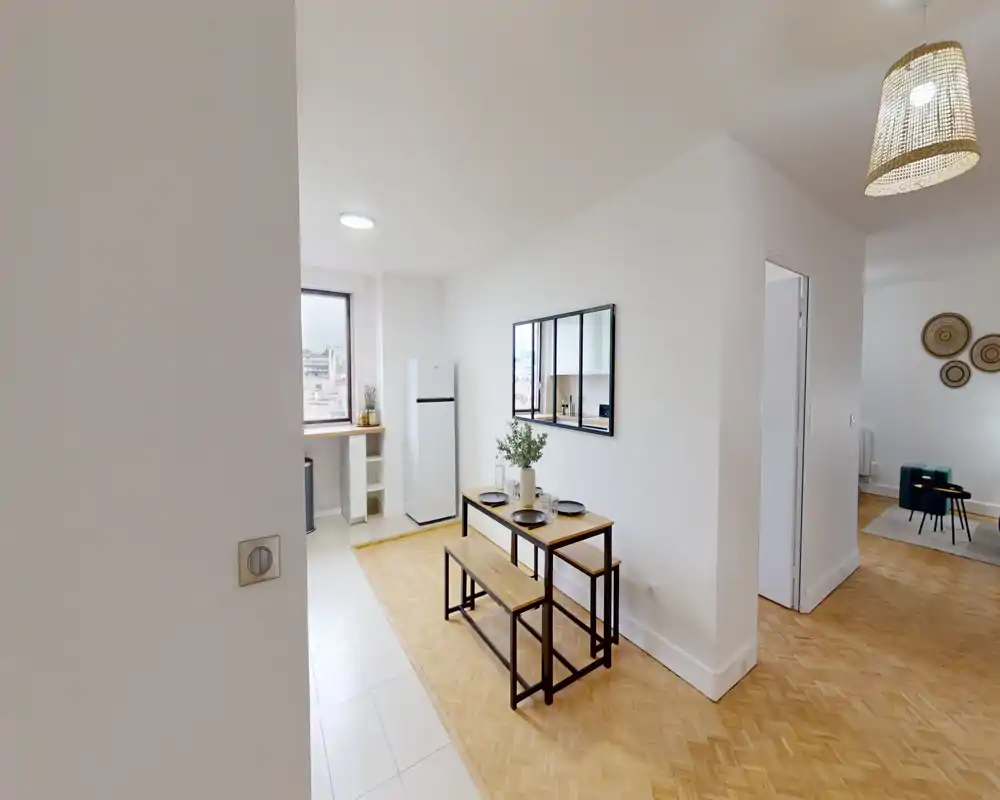 Bright, modern dining nook with a wooden table and bench seating, styled with dishware and a small plant; the space opens toward the kitchen and living area.