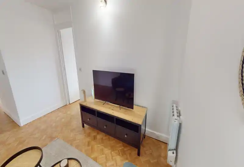Simple, well-lit living area with a TV on a console, parquet flooring and a neutral, minimal decor—suitable for a compact lounge or studio setup.