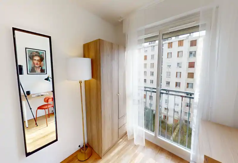 Coin de chambre lumineux et minimaliste avec une armoire, une lampe sur pied et une grande fenêtre ; un miroir haut reflète une petite partie de la zone adjacente. La pièce est aérée et bien éclairée, adaptée aux annonces mettant en avant la lumière naturelle et le rangement.