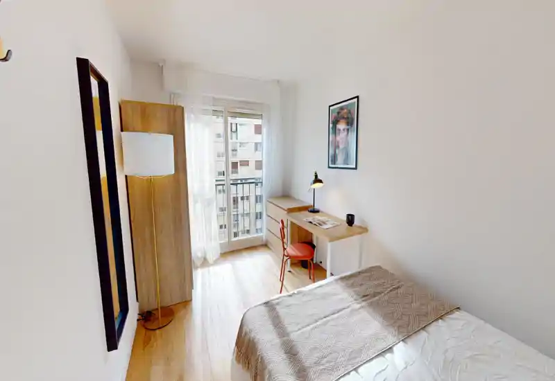 Chambre simple lumineuse avec lit bien fait, sol en bois, bureau et fenêtre donnant sur balcon — idéale pour étudiants ou professionnels recherchant un espace compact et lumineux.