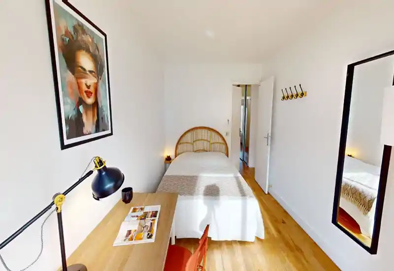 Chambre lumineuse et étroite avec lit simple et tête de lit en rotin, bureau et grand miroir mural — décoration minimaliste, sol en bois chaleureux offrant une atmosphère cosy et bien éclairée, idéale pour une personne.