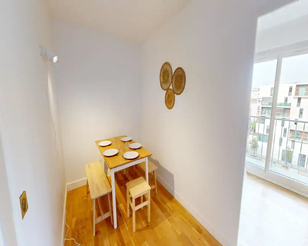 Coin repas compact avec une petite table en bois, bancs et tabourets dressés pour quatre personnes. La lumière naturelle venant du balcon et le sol en bois chaleureux créent un espace repas minimaliste et accueillant.