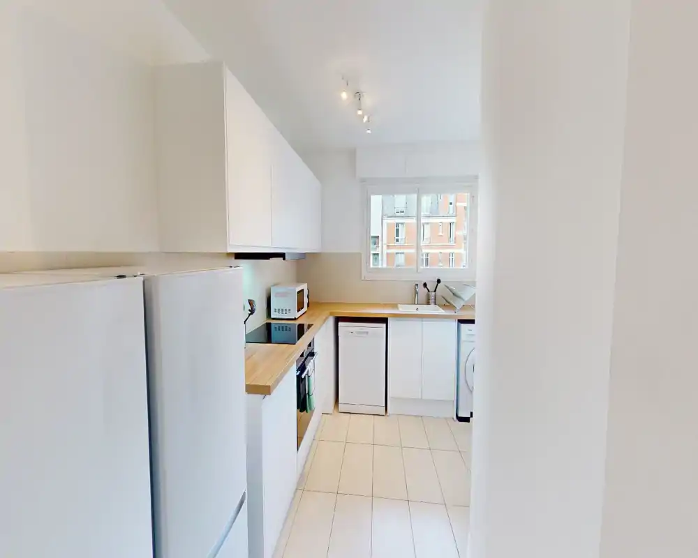 Cuisine moderne et lumineuse avec meubles blancs, plan de travail en bois, four encastré, lave-vaisselle et fenêtre apportant la lumière naturelle — agencement compact et soigné adapté aux appartements urbains.