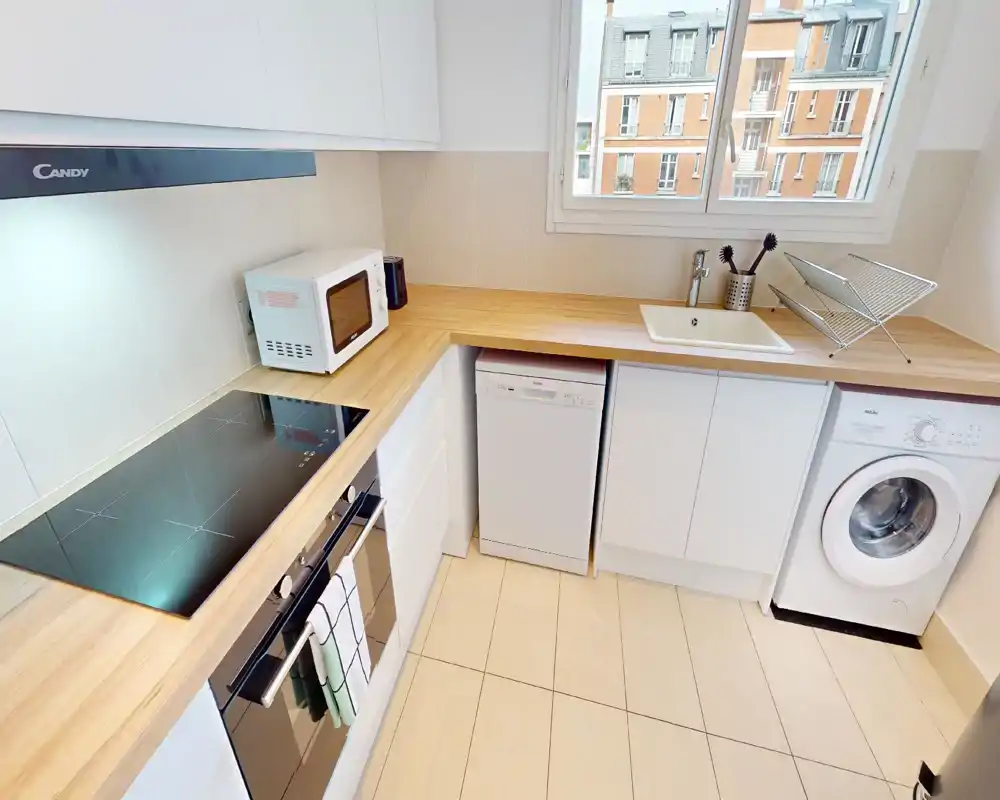 Cuisine moderne équipée avec plans de travail en bois, plaque à induction, four, lave-vaisselle et lave-linge intégrés; fenêtre lumineuse et rangement soigné.