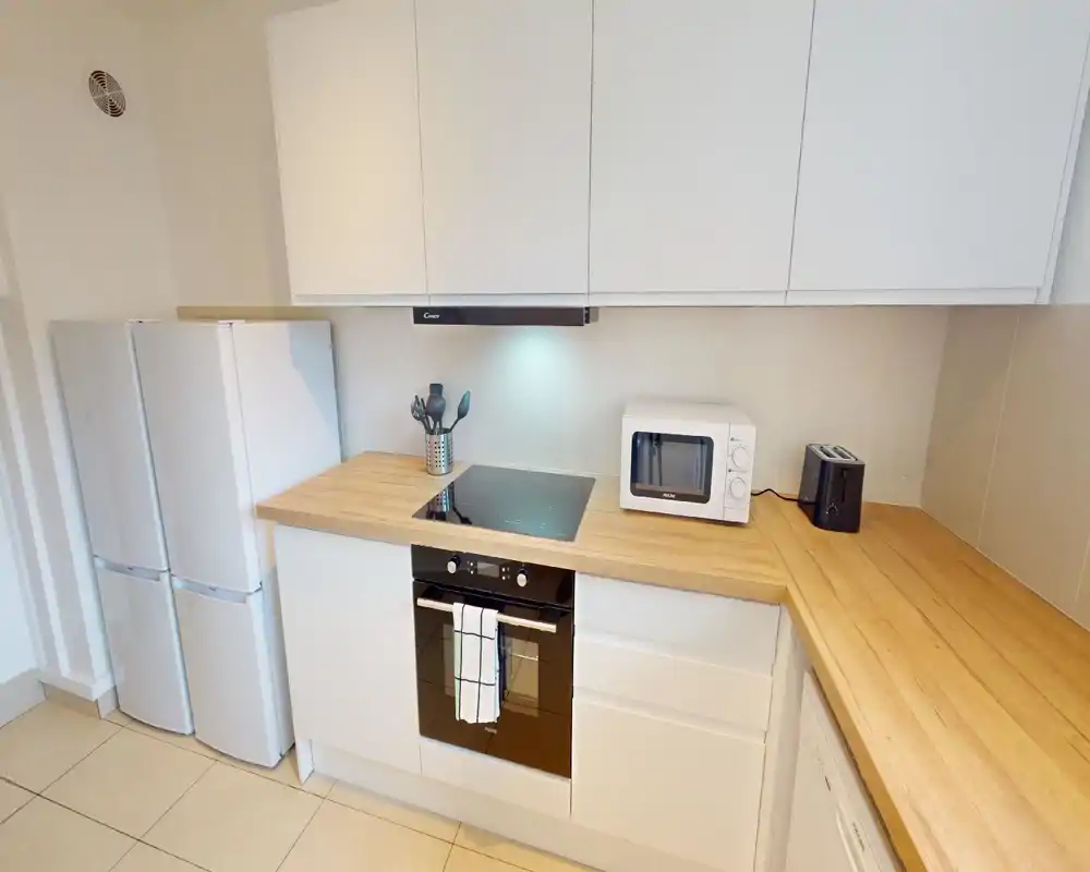 Cuisine moderne et lumineuse avec meubles blancs, plans de travail en bois, plaque à induction et four encastré ; micro-ondes, grille-pain et double réfrigérateur visibles — espace propre et fonctionnel pour location courte durée.
