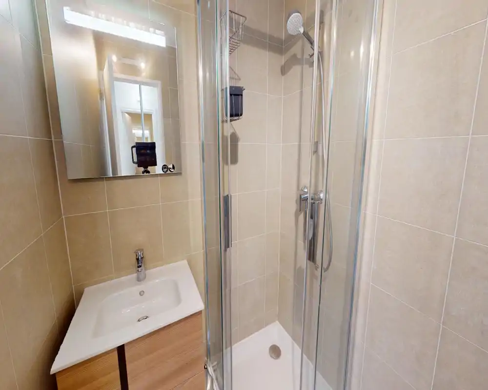 Salle de bains moderne et compacte avec cabine de douche en verre, robinetterie chromée et meuble vasque; carreaux beige neutre pour un aspect propre.