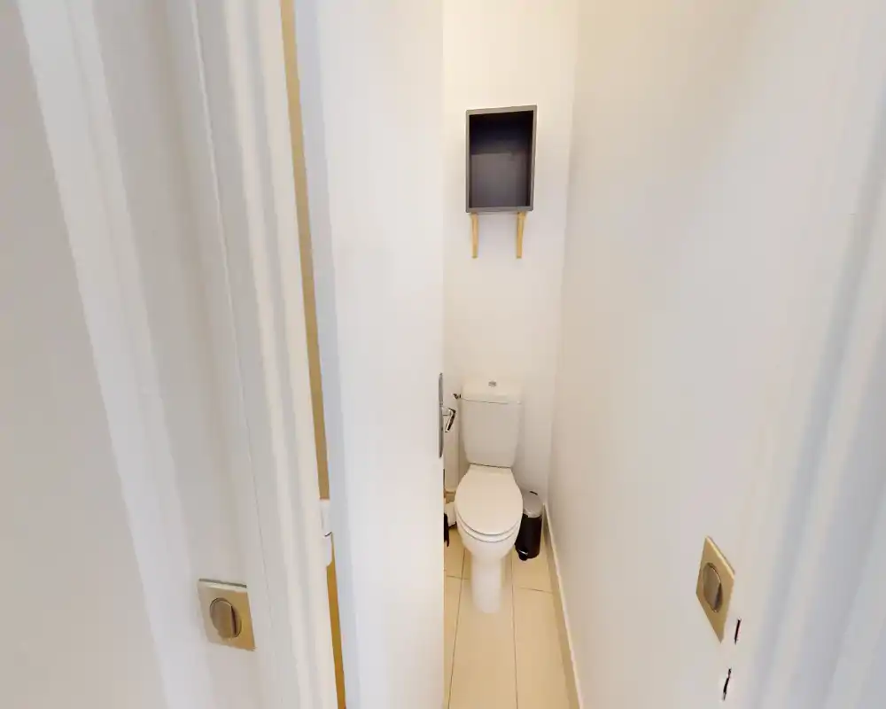 Toilettes compactes et propres avec WC standard, sol en carrelage clair et une petite étagère encastrée au-dessus pour rangement.