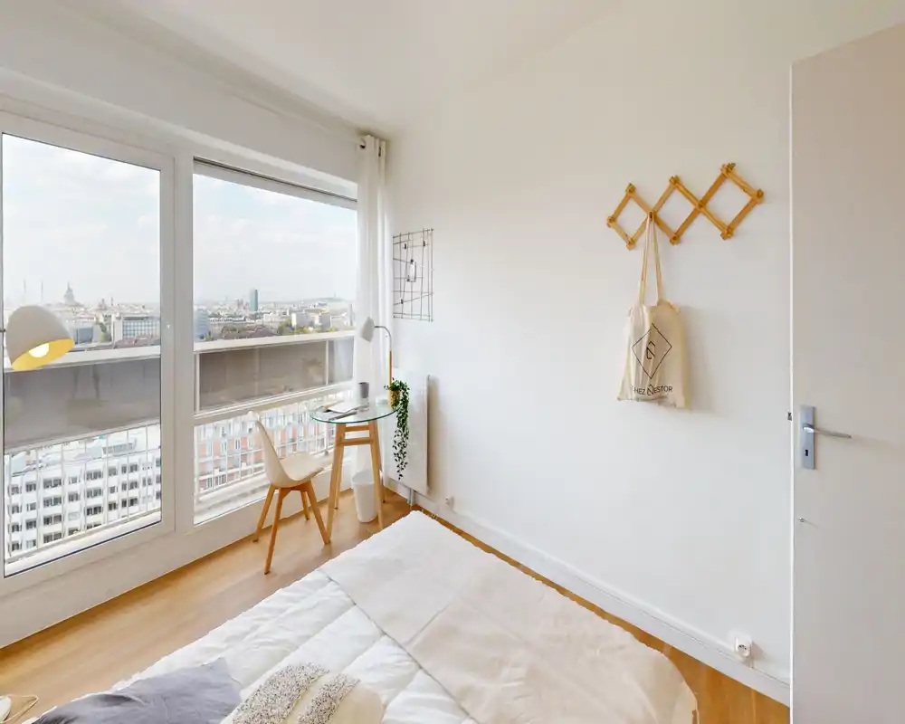 Chambre lumineuse et accueillante avec grande fenêtre offrant une vue sur la ville, lit bien fait et un petit espace bureau près de la fenêtre.