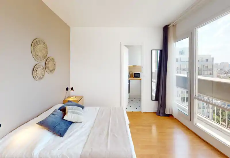 Chambre lumineuse et moderne avec grande fenêtre et accès au balcon ; lit bien fait avec coussins et sol en bois chaleureux pour une ambiance accueillante.