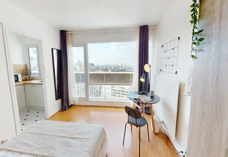 Chambre moderne et lumineuse avec grande fenêtre donnant sur la ville, lit bien fait et petit bureau près du radiateur — idéale pour travailler et se détendre.