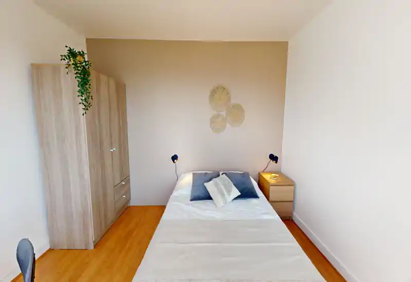 Chambre lumineuse et soignée avec lit central, table de chevet et armoire ; parquet chaud et décoration murale discrète créent une ambiance calme et accueillante.