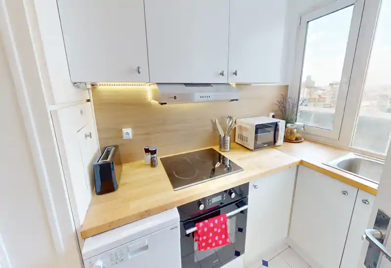 Cuisine compacte et moderne avec plan de travail en bois, plaque électrique, four encastré, micro-ondes et évier près d'une fenêtre lumineuse — idéale pour un appartement en ville.