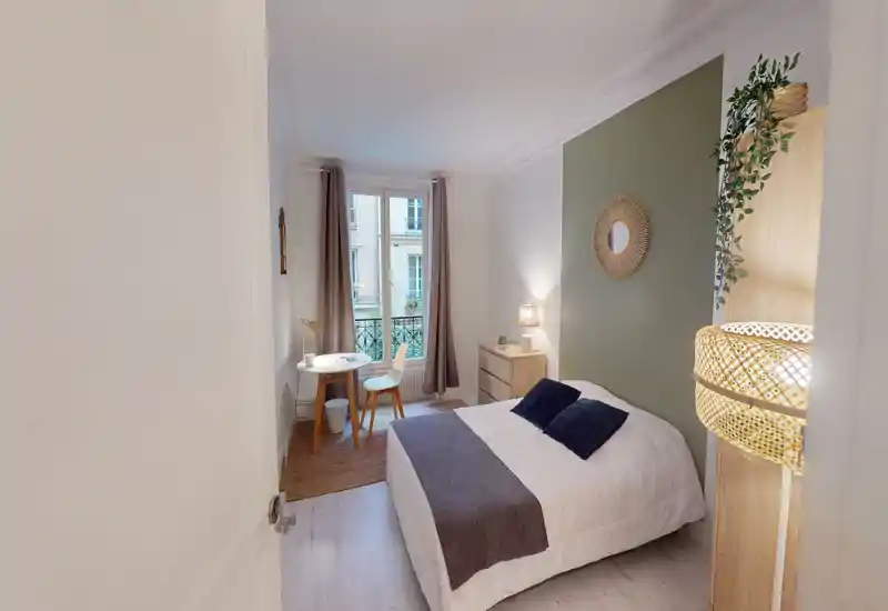 Bright, stylish bedroom with a double bed, soft textiles, bedside lamp and a small workspace by the window. Large window with curtains provides natural light; tasteful green accent wall and rattan details add warmth.