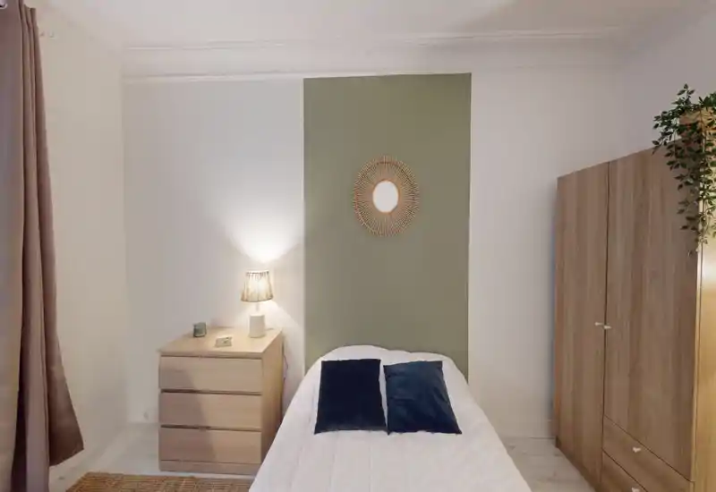 Bright, neatly styled single bedroom with a central bed, bedside chest, warm lamp and wardrobe — calm neutral tones and decorative mirror accent.