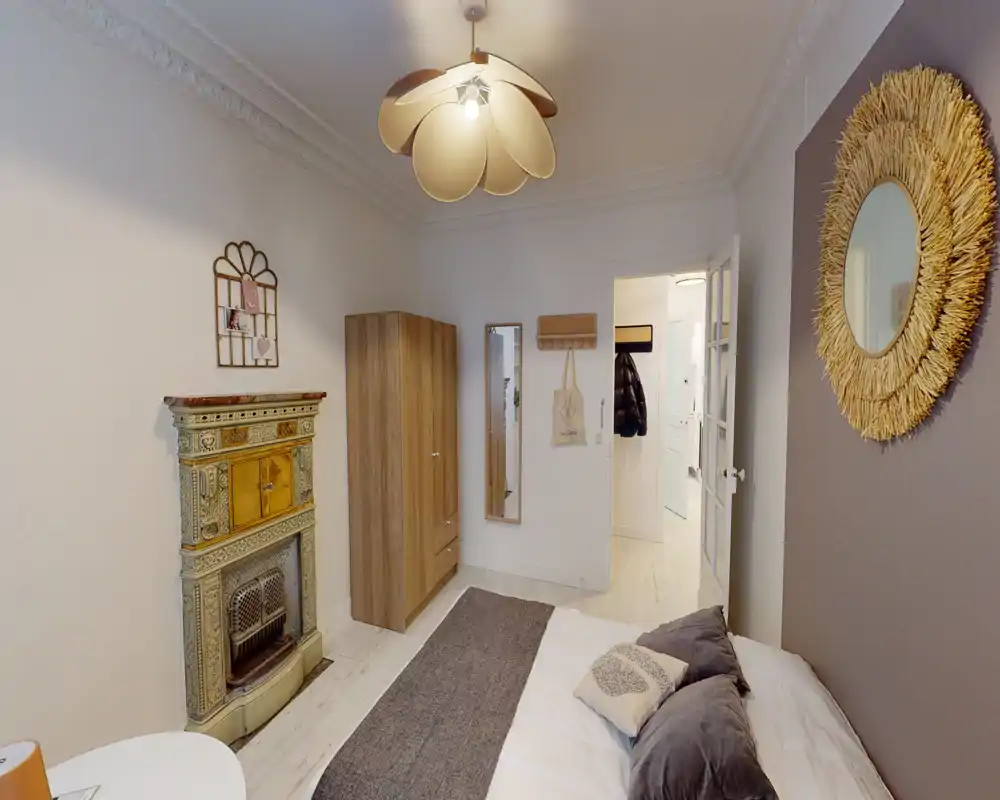 Bright, stylish bedroom with a comfortable double bed, decorative pillows, a rug and tasteful wall decor including a round mirror and vintage fireplace.