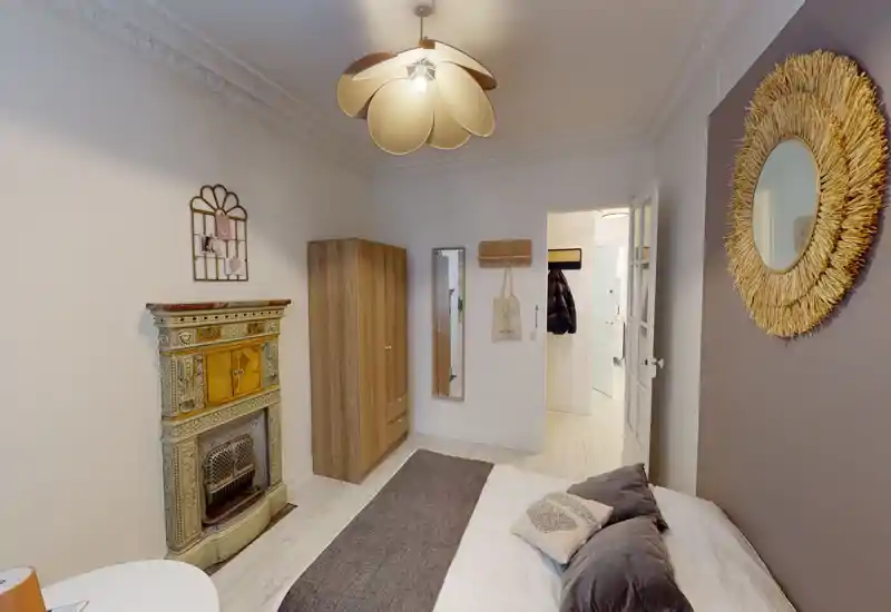 Bright, stylish bedroom with a comfortable double bed, decorative pillows, a rug and tasteful wall decor including a round mirror and vintage fireplace.