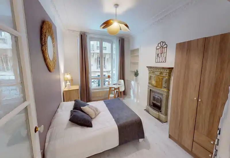 Bright, stylish double bedroom with a neatly made bed, decorative fireplace, wardrobe and a small desk by the window — ideal for comfortable stays in a central city apartment.