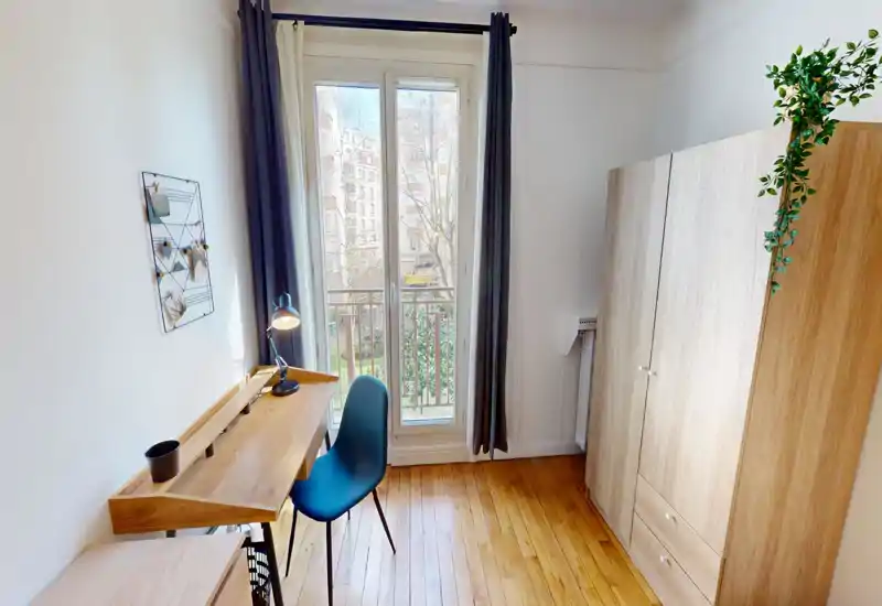 Angolo camera luminoso e arredato in modo semplice con una grande finestra, armadio in legno e una scrivania vicino alla finestra perfetta per lavorare. Le tende chiare e la piantina aggiungono un tocco casalingo.