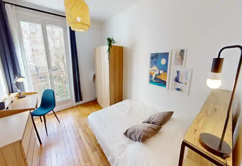 Camera da letto luminosa e ben allestita con letto matrimoniale, pavimento in legno e ampia finestra che dona molta luce naturale. Arredo moderno e minimale con quadri, armadio e scrivania — perfetta per annunci di affitto.