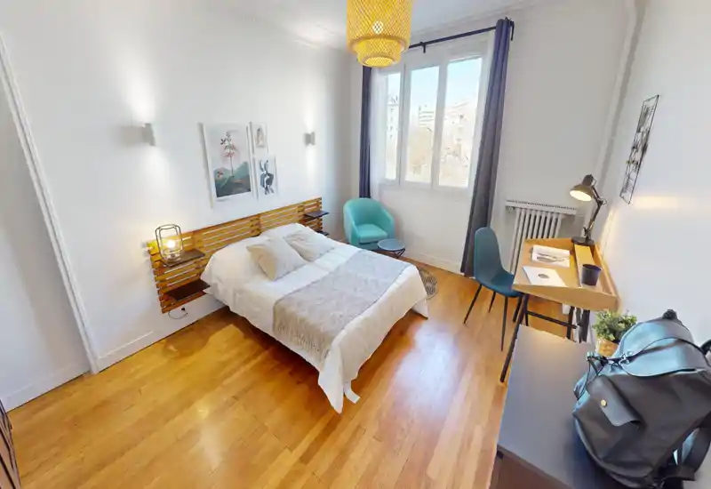 Bright, modern bedroom with a centrally placed double bed, cozy textiles and a small workspace by the window. Natural light from a large window, wooden floors and calming neutral decor create a welcoming atmosphere.