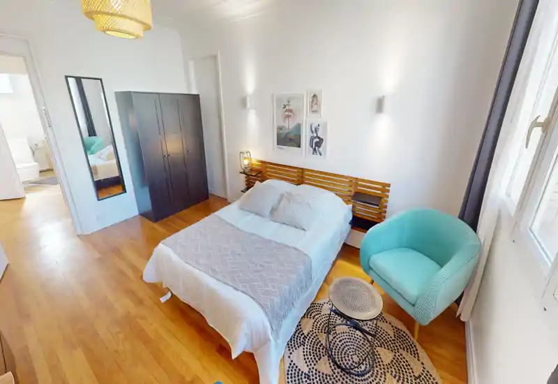 Bright, stylish bedroom with a centrally placed double bed, wooden headboard, wardrobe, and a turquoise armchair by the window — well lit and tastefully decorated.