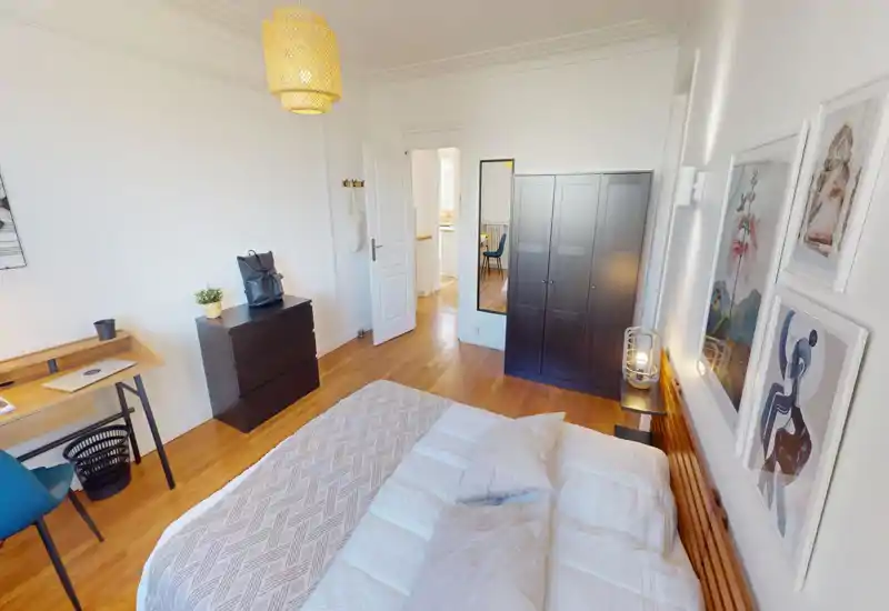 Bright, stylish bedroom with a large bed in the foreground, wooden floor, dark wardrobe and desk area — cozy linens and framed artwork create a welcoming atmosphere.