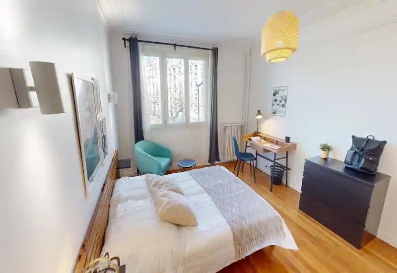 Bright, modern bedroom with a large bed, wooden floor, reading nook by the window and a compact work desk. Natural light fills the room through a big window, creating a warm and inviting atmosphere.