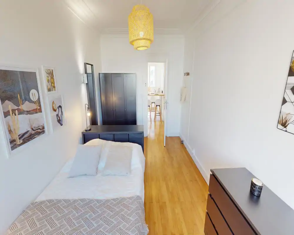 Camera da letto luminosa e ben arredata con un letto matrimoniale in primo piano, pavimenti in legno caldi, mobili contenitori scuri e quadri decorativi. Una grande porta si apre verso la zona pranzo/cucina luminosa, creando un senso di ampiezza.