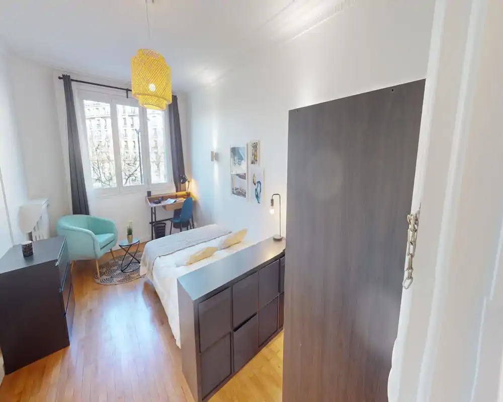 Camera da letto luminosa e arredata con letto matrimoniale, lampade da comodino, una scrivania vicino alla finestra e una poltrona confortevole. La grande finestra regala molta luce naturale e il pavimento in legno rende l’ambiente accogliente.