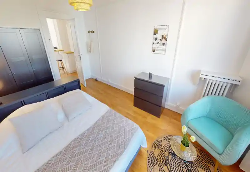 Camera da letto luminosa e curata con un ampio letto, cassettiera e un angolo relax con poltrona turchese e tappeto decorativo.