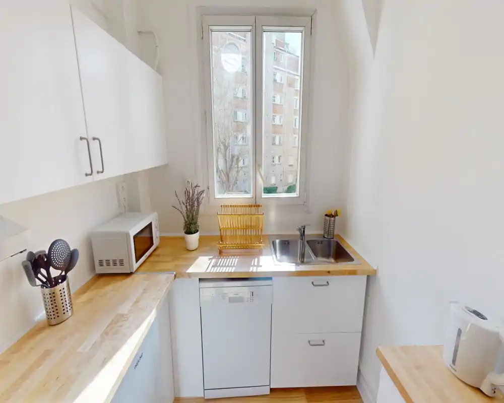 Cucina compatta e luminosa con piano in legno, lavello integrato, lavastoviglie e microonde. La luce naturale dalla finestra centrale valorizza le superfici ordinate e gli elementi di arredo moderni.