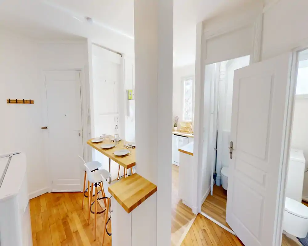 Cucina luminosa a vista con bancone in legno apparecchiato, mobili bianchi e caldo pavimento in parquet — uno spazio compatto e accogliente perfetto per pasti e incontri informali.