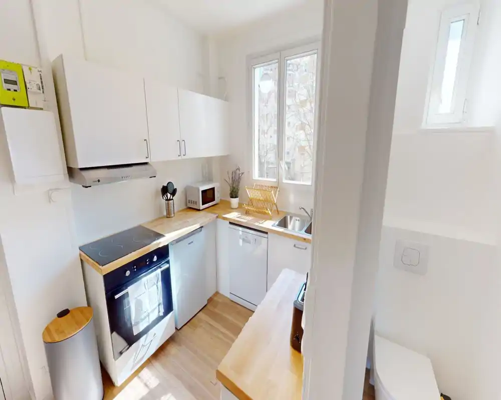 Cucina compatta e luminosa con pensili bianchi, piani in legno, piano cottura elettrico, lavastoviglie e lavello vicino alla finestra — perfetta per un piccolo appartamento.